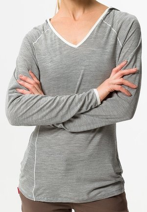 Persona che indossa una maglietta a maniche lunghe a righe sottili con scollo a V, con le braccia incrociate sul petto, su uno sfondo chiaro uniforme.