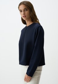 Top blu navy a maniche lunghe realizzato in tessuto testurizzato, con scollatura rotonda e linee pulite, abbinato a pantaloni bianchi aderenti.