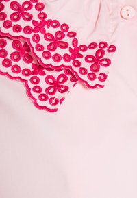 Tissu rose avec bord festonné orné d'une broderie en dentelle florale rose vif et un bouton rose pâle près du col.