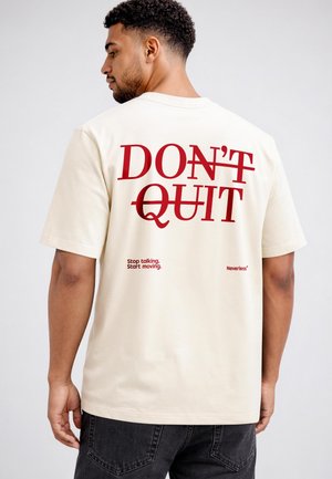 Mann mit kurzen lockigen Haaren, der ein weißes T-Shirt mit rotem Text "DON'T QUIT" und "Stop talking. Start moving." auf dem Rücken trägt.