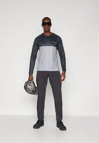 Langermet, totonet sykkeltrøye i grått og svart, med logo og hjelm i hånden, kombinert med mørke, lette bukser og svarte joggesko.