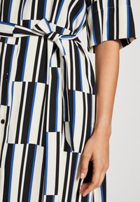 Abito a camisole a righe in nero, bianco e blu. Presenta maniche corte, una cintura in vita e dettagli con bottoni. Tessuto liscio con motivi geometrici.