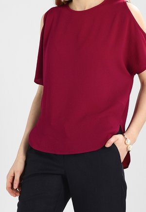 Kobieta w bordowym topie bez rękawów z wycięciami na ramionach i czarnych spodniach, z ręką w kieszeni, na tle jednolitego szarego tła.