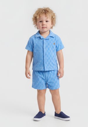 Νήπιο με ξανθά, σγουρά μαλλιά φορά μπλε πουκάμισο με κοντά μανίκια και κουμπιά και ταιριαστό σορτς, στέκεται με μπλε ναυτικά παπούτσια χωρίς κορδόνια.