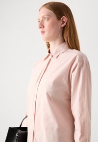 Light pink button-up shirt with a collar, long sleeves, and a subtle embroidered logo. Smooth fabric and relaxed fit, paired with a black handbag.