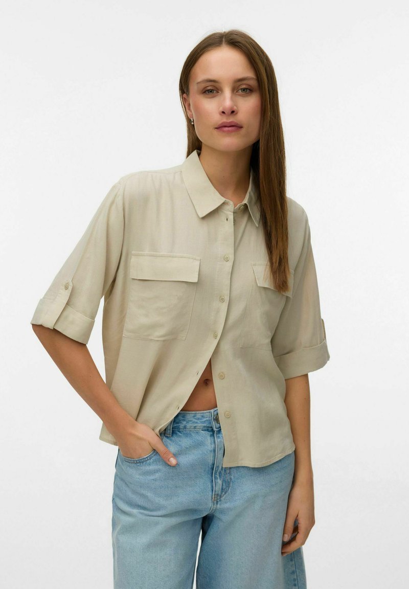Femme aux cheveux longs, raides et bruns, portant une chemise beige à boutons avec les manches retroussées et un jean bleu clair, debout devant un fond blanc uni.