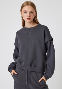 Sudadera gris de gran tamaño con puños y dobladillo acanalados, que presenta un detalle de punto de cruz en el cuello, combinada con pantalones de chándal a juego.