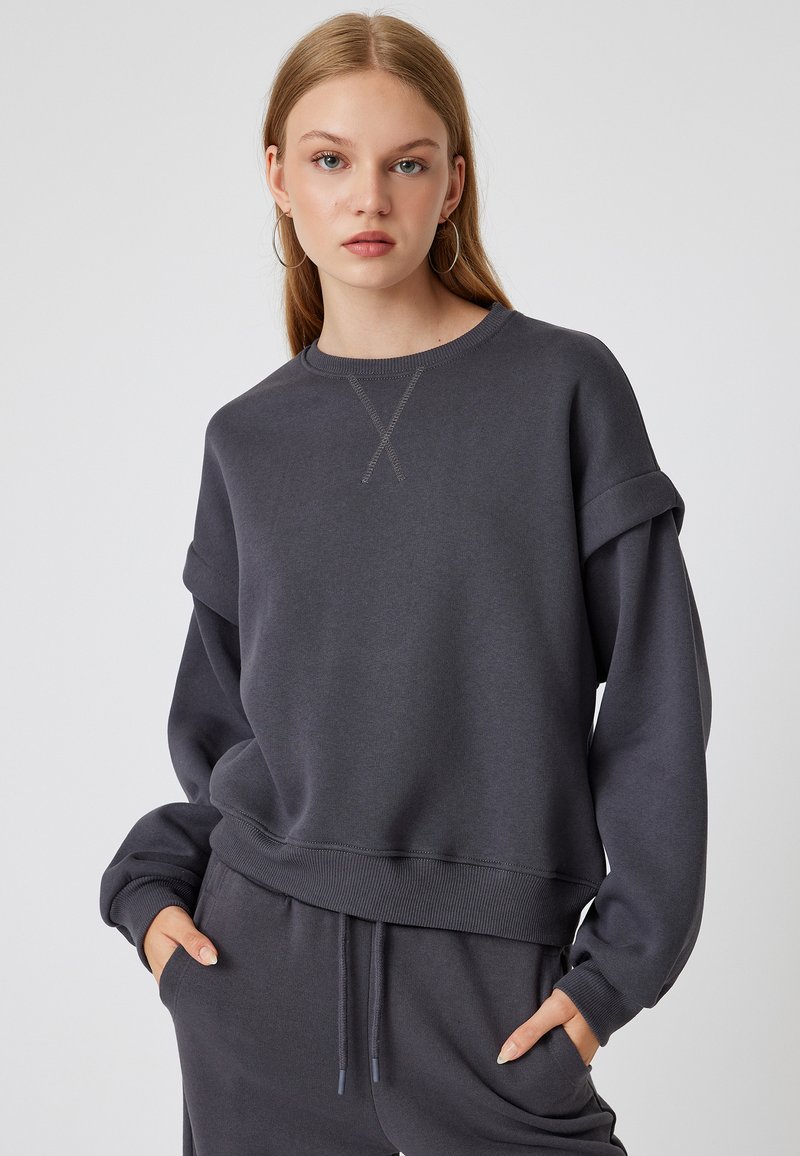 Sudadera gris de gran tamaño con puños y dobladillo acanalados, que presenta un detalle de punto de cruz en el cuello, combinada con pantalones de chándal a juego.
