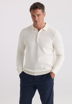 Homme portant un polo texturé blanc cassé à manches longues et un pantalon foncé, les mains dans les poches, devant un fond uni.