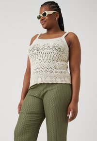 Cream crocheted tank top with intricate patterns, paired with textured olive-green pants. Clear sunglasses complete the look.