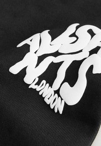 Melns audums ar izteiksmīgu baltu tekstu "ALSA NYTS LONDON" izplūdušā, viļņotā fontā, uzdrukātu uz virsmas.