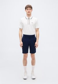 Πόλο πουκάμισο σε λευκό με μαύρες λεπτομέρειες, γιακάς με φερμουάρ, συνδυασμένο με σορτς σε ναυτικό μπλε. Το μοντέλο φοράει λευκά αθλητικά παπούτσια και ανοιχτόχρωμες κάλτσες. Μινιμαλιστικός σχεδιασμός.