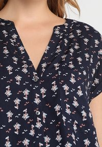 Marinblus med V-hals, korta ärmar och knappfront. Har blommiga och geometriska mönster i korall och vitt. Mjuk tygtextur.