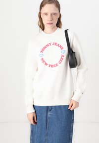 Hvit sweatshirt med "TOMMY JEANS NEW YORK CITY" i rosa og blå skrift, kombinert med et denimskjørt og en svart skulderbag.