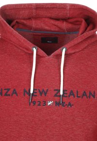 Rode hoodie met textielstructuur, voorzien van een trekkoordkap, geborduurde tekst "NZ New Zealand 1923 NZA" en een klein vlagdetail.