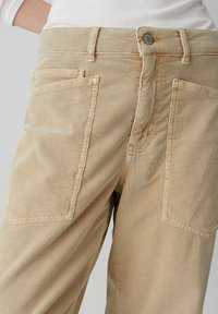 Pantaloni in corduroy beige con ribbatura verticale, due tasche posteriori e chiusura con bottone in vita. Dettagli di cucitura discreti visibili.