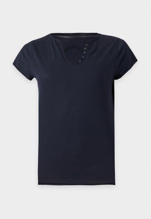 Femme debout portant une chemise noire à manches courtes et un pantalon large en cuir noir, une main dans la poche, sur fond blanc.