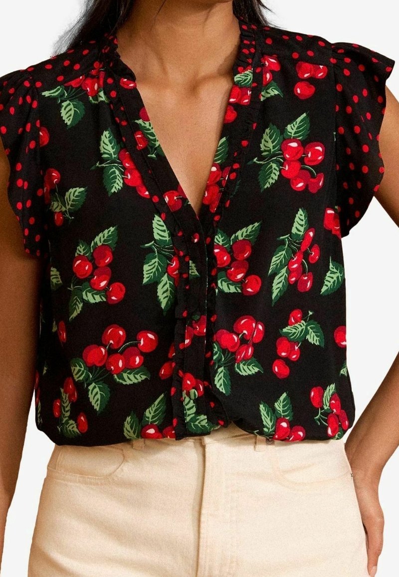 Femme portant un chemisier noir sans manches avec un imprimé cerise rouge et feuilles vertes, associé à un pantalon beige clair.