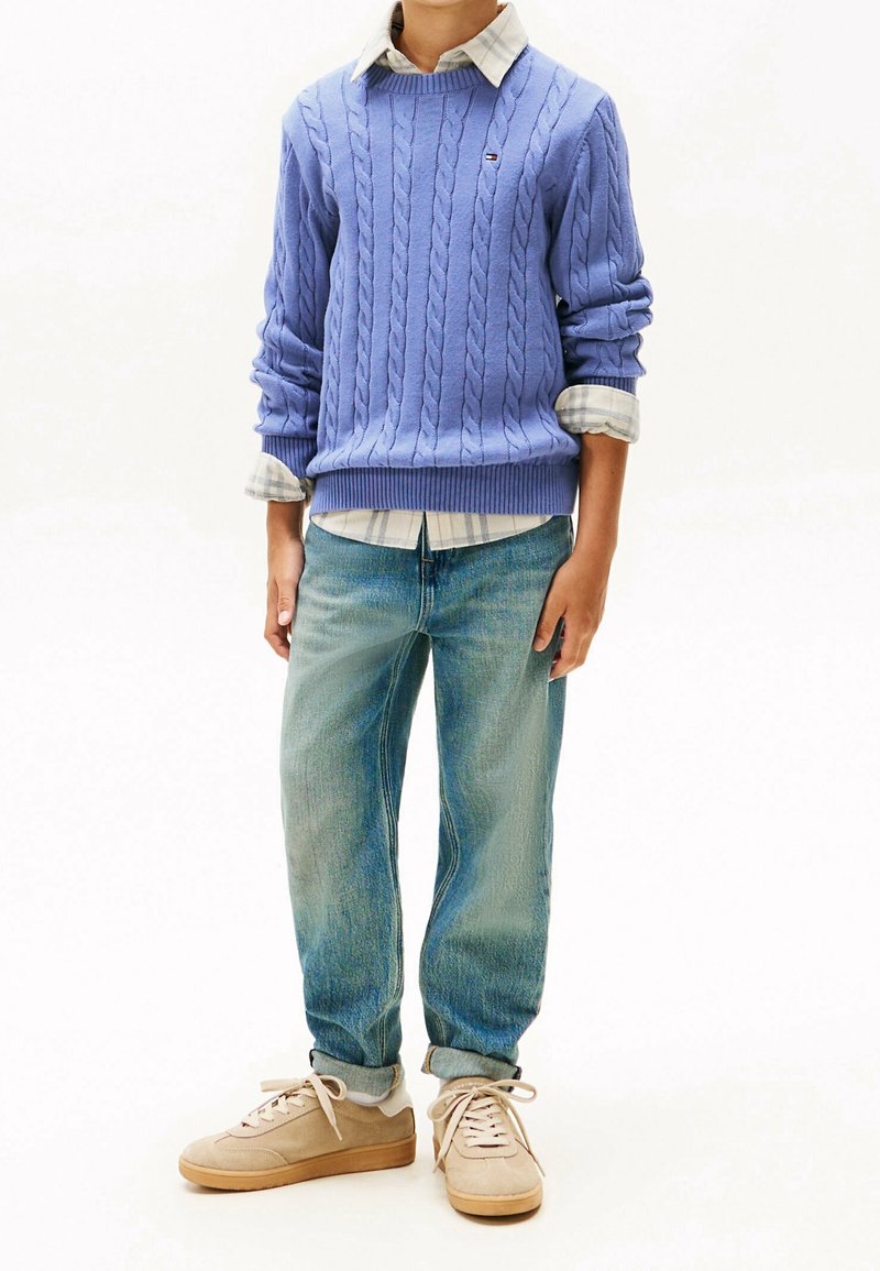 Enfant portant un pull bleu clair à torsades par-dessus une chemise à carreaux, un jean bleu ample retroussé aux chevilles et des baskets beiges à lacets.