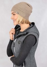 Gebreide beige muts met geribbelde structuur, gedragen met een grijs mouwloos vest voorzien van een zwart logo en een witte rits.