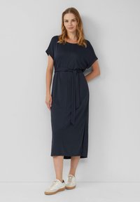 Marineblaues, knielanges Kleid aus weichem Stoff mit kurzen Ärmeln, tailliertem Gürtel und einem Seitenschlitz. Trägt dazu weiße Sneakers.