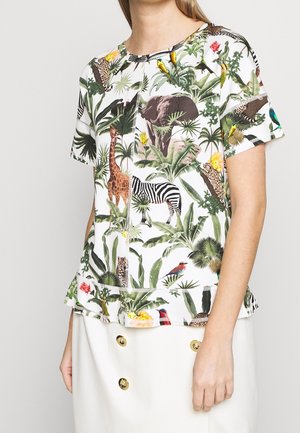 Kobieta ubrana w białą bluzkę z krótkim rękawem z kolorowym nadrukiem zwierząt i roślin dżungli, w zestawie z białą spódnicą z złotymi guzikami.