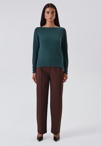 Vrouw staat in een donkergroene sweater met lange mouwen en een broek met wijde pijpen in bruin, met zwarte puntige schoenen tegen een effen achtergrond.