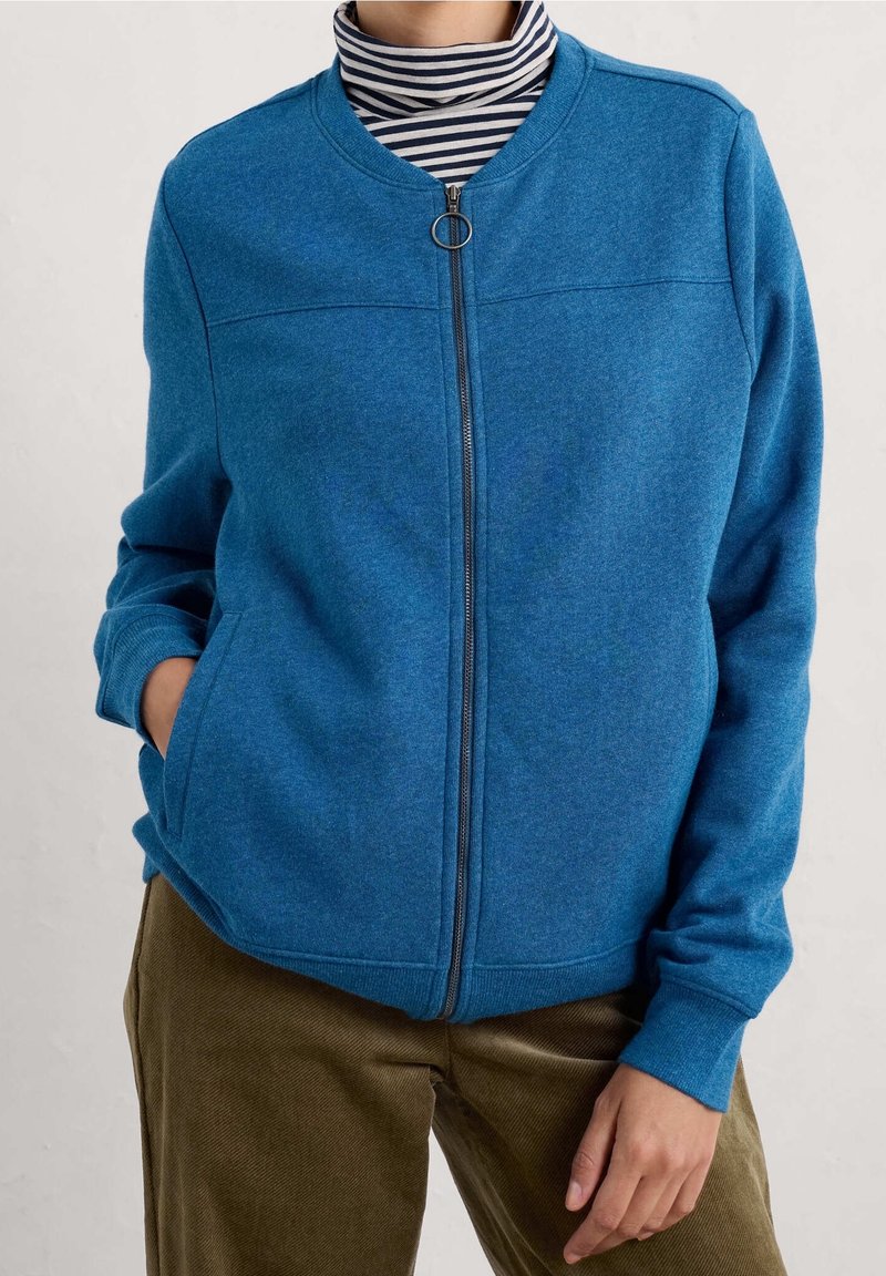 Personne portant une veste zippée bleue par-dessus un col roulé rayé noir et blanc et un pantalon marron, une main dans la poche de la veste.