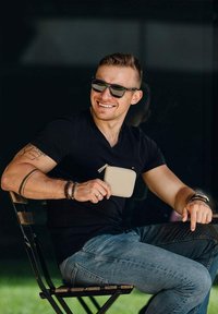Béžová peněženka na zip držena v ruce, nošena s černým tričkem s výstřihem do V a džínami, sedící na dřevěné židli venku.