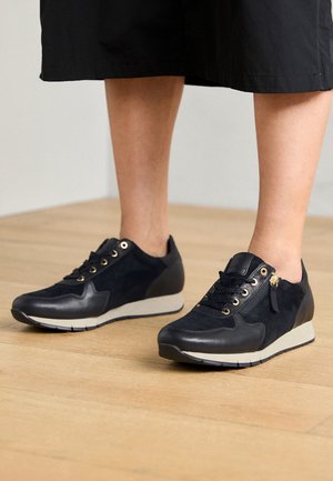 Osoba nosząca czarne skórzane i zamszowe sneakersy ze złotymi oczkami i bocznym zamkiem błyskawicznym, stojąca na drewnianej podłodze.