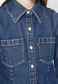 Koszula dżinsowa z kołnierzykiem w szpic, dwoma kieszeniami na piersi oraz kontrastującymi guzikami. Kolor ciemnoniebieski z fakturowanym wykończeniem. Obecna czerwona metka z logo.
