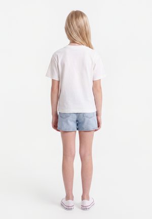 Ung person med langt blondt hår står med ryggen til iført en hvid kortærmet skjorte, lyseblå denimshorts og hvide sneakers.