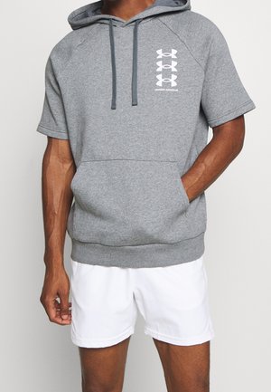 Mann trägt einen grauen Under Armour Hoodie mit kurzen Ärmeln und frontalem Taschen, sowie weiße Sportshorts und steht vor einem schlichten Hintergrund.