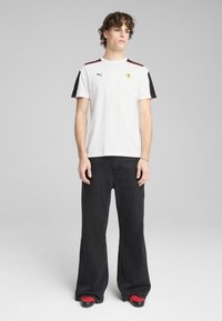 Camiseta blanca con acentos en las mangas en negro, que presenta un logo de Ferrari en amarillo y una franjas rojas. Combinada con unos vaqueros de pierna ancha negros y zapatos rojos.