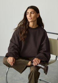 Eine Frau mit langen, dunklen Haaren sitzt im Schneidersitz auf einem Stuhl, trägt einen lockeren, dunkelbraunen Pullover, olivfarbene Hosen und dunkelbraune Stiefel.
