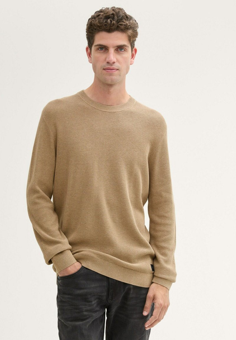 Beige Waffelstrick-Pullover mit langen Ärmeln, Rippkragen und Rippbündchen, kombiniert mit dunklen Jeans. Glatte Textur, lockere Passform.