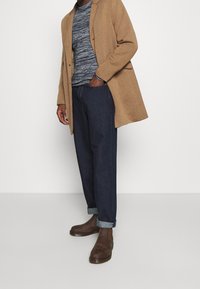 Brązowy płaszcz z mieszanki wełny na granatowym swetrze w paski, ciemnoniebieskie dżinsy oraz brązowe skórzane buty z podwiniętymi mankietami.