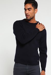 Pull en tricot bleu marine avec un col rond, des poignets et un ourlet côtelés. Texture douce, ajusté à la taille, porté avec un jean foncé.