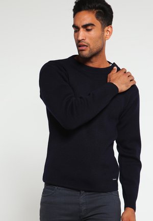 Maglione in maglia blu navy con scollo rotondo, polsi e orlo a coste. Tessuto morbido, aderente in vita, indossato con jeans scuri.