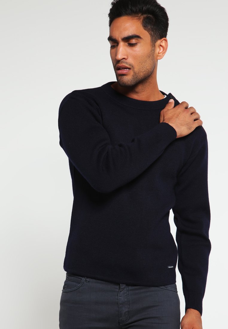 Pull en tricot bleu marine avec un col rond, des poignets et un ourlet côtelés. Texture douce, ajusté à la taille, porté avec un jean foncé.
