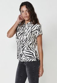 Camisa de botón de manga corta en tejido ligero con un patrón de rayas de cebra en blanco y negro. Incluye dos bolsillos en el pecho.