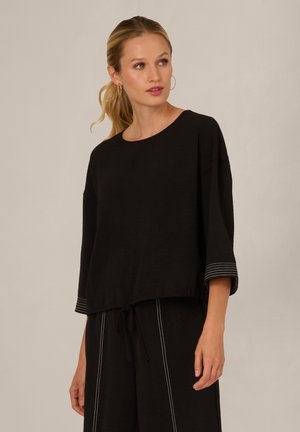 Woman with blonde hair in a ponytail wearing black loose top with three-quarter sleeves and matching black pants with white stitch details.