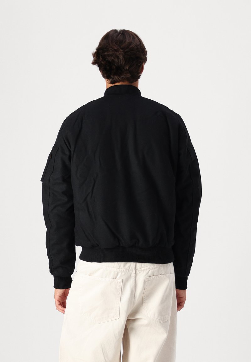 Schwarze Bomberjacke mit gerippten Bündchen und Kragen, seitlicher Tasche, getragen über hellen Hosen. Rückansicht, glatte Textur.