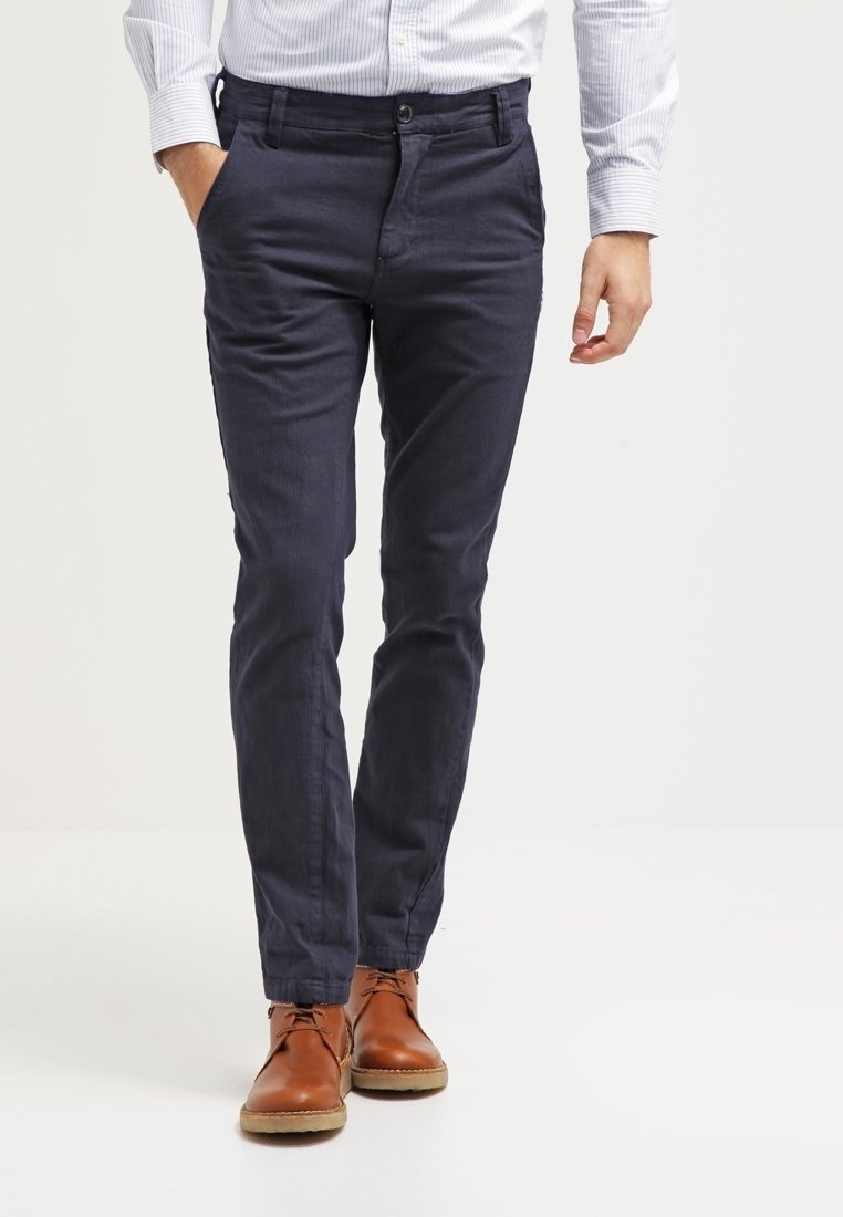 Mann trägt marineblaue, schmal geschnittene Hose, hellblau gestreiftes Hemd und braune Leder-Schnürschuhe, steht mit einer Hand in der Tasche.