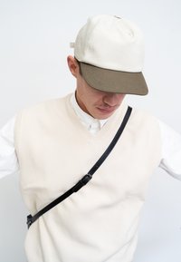 Beige Kappe mit einem dunkelgrünen Schirm, strukturierter Stoff, getragen mit einem beigefarbenen ärmellosen Pullover und einem schwarzen Umhängegurt.