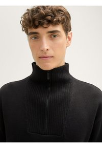 Schwarzer Reißverschluss-Pullover mit hohem, geripptem Kragen und strukturiertem Design auf der Vorderseite. Aus einem glatten, weichen Material gefertigt.