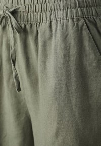 Zielone lniane spodnie z elastycznym paskiem i zawiązywanym sznurkiem, ukazujące fakturę i detale kieszeni.