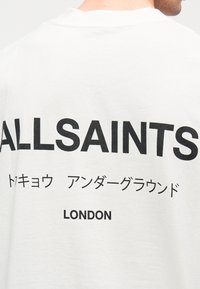 Λευκό βαμβακερό T-shirt με έντονα μαύρα γράμματα στην πλάτη: "ALLSAINTS", ιαπωνικούς χαρακτήρες και "LONDON" στο κάτω μέρος.