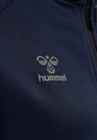 Nahaufnahme von marineblauem Stoff mit einem hellgrauen stilisierten Bienenlogo und dem Wort „hummel“ darunter.