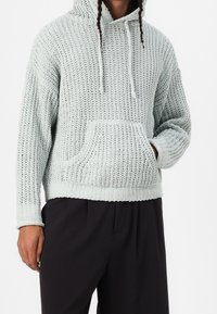 Sudadera de punto verde menta claro con un corte holgado, patrón texturizado, cordones y un bolsillo delantero; llevada sobre pantalones negros.